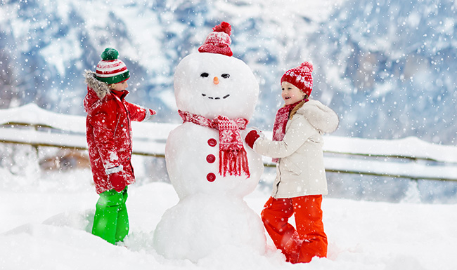 Snögubbe med röd mössa och halsduk, två barn i vinterkläder leker i snön.