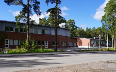 Skolbyggnad i två plan, tegelfasad med grå övre del, tallar, flaggstång och blå himmel.