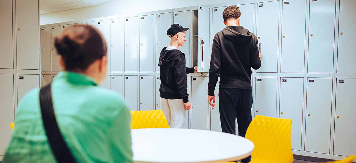 Tonåringar i skolkorridor med ljus elevskåp. En flicka sitter med ryggen mot kameran i grön blus, två pojkar står vid skåpen i svarta hoodies.