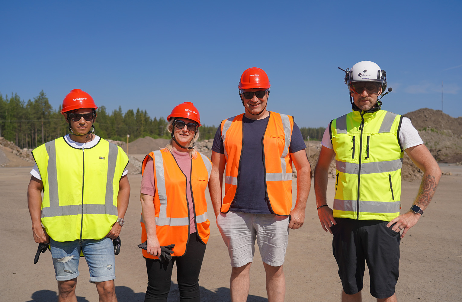 Fyra personer står på en sandplan, blå himmel. De bär reflexvästar och skyddshjälmar.