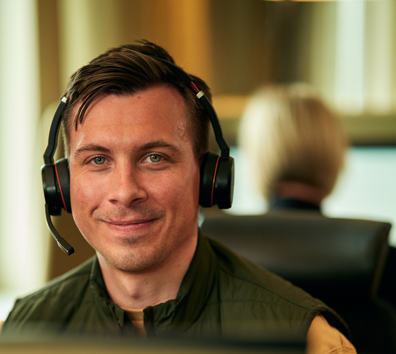 Leende manlig kundservicemedarbetare med mörkt hår, headset och grön täckväst och gul tröja i kontorsmiljö.
